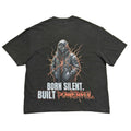 A dark gray graphic t-shirt laid flat on a light gray background, showing the back side with a detailed print of a hooded figure and the text 'BORN SILENT. BUILT POWERFUL'.