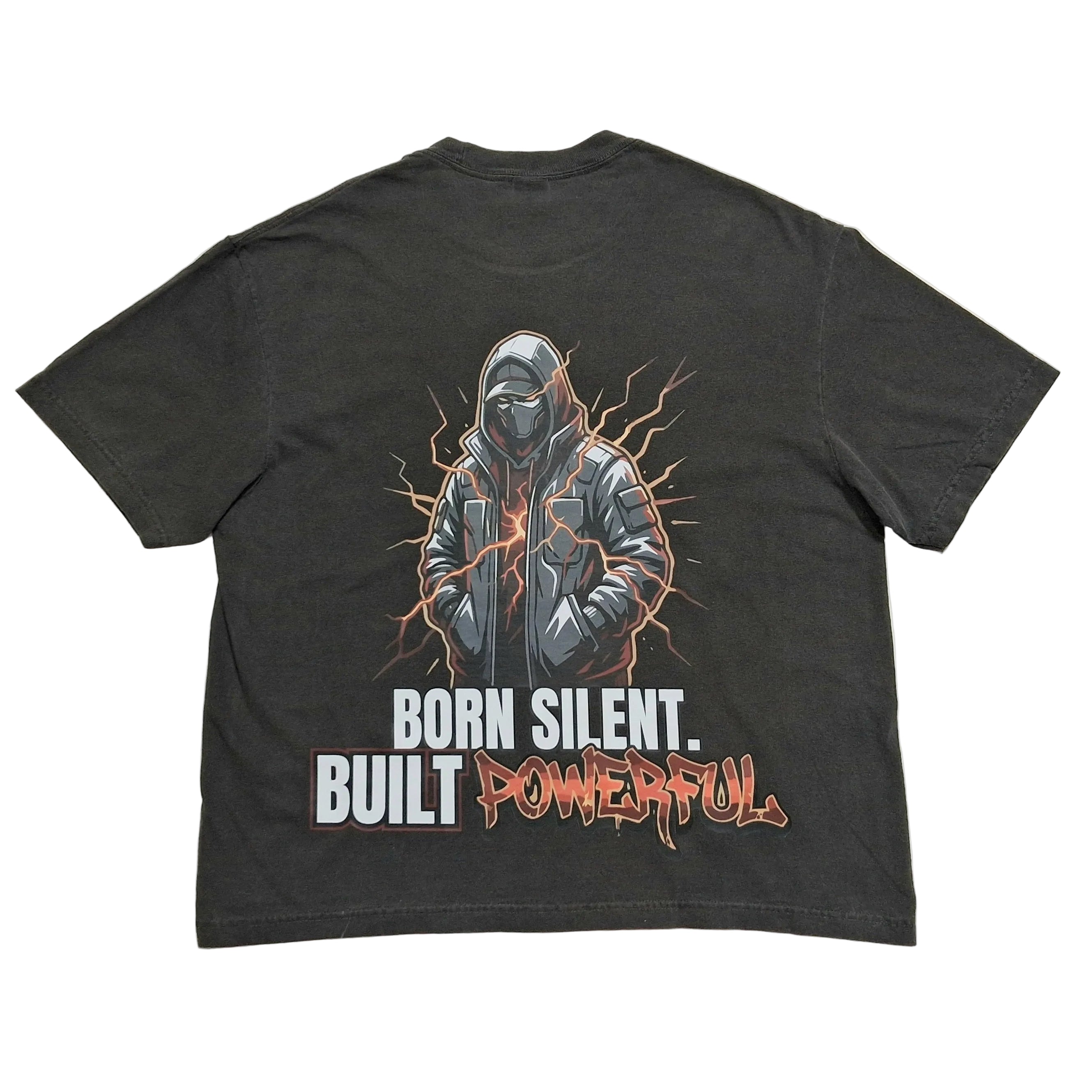A dark gray graphic t-shirt laid flat on a light gray background, showing the back side with a detailed print of a hooded figure and the text 'BORN SILENT. BUILT POWERFUL'.