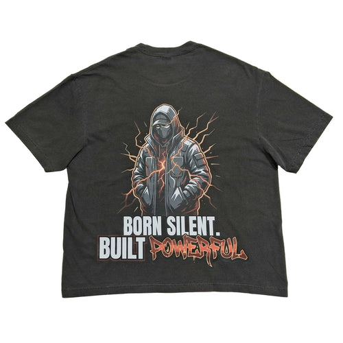 A dark gray graphic t-shirt laid flat on a light gray background, showing the back side with a detailed print of a hooded figure and the text 'BORN SILENT. BUILT POWERFUL'.