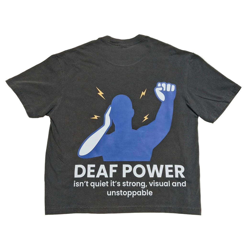 A dark gray graphic t-shirt laid flat on a light beige background. The back of the shirt features a blue silhouette of a person with a raised fist, yellow lightning bolts, and white text that reads 'DEAF POWER isn't quiet it's strong, visual and unstoppable'.