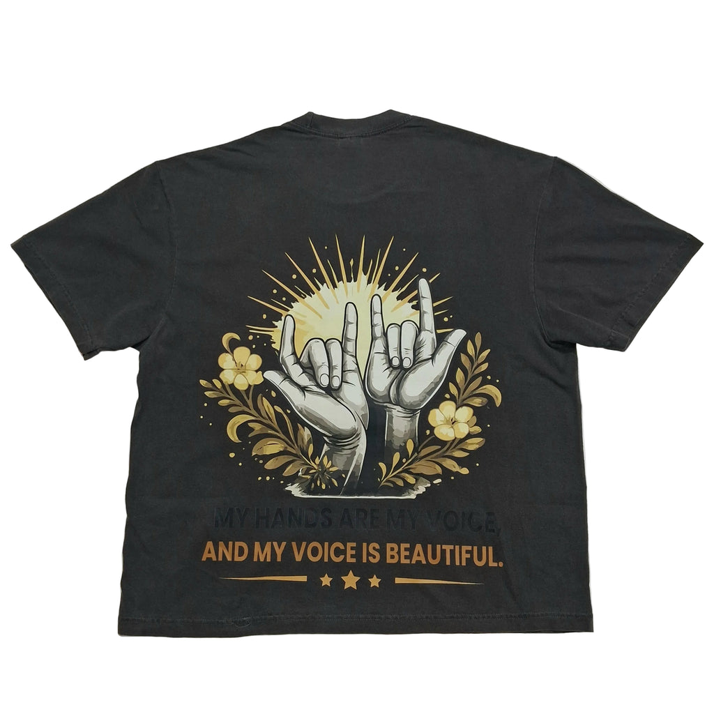 A black short-sleeve t-shirt laid flat against a light gray background, showing the back side with a graphic print of two hands making the shaka sign, surrounded by yellow flowers and rays, with the text 'MY HANDS ARE MY VOICE AND MY VOICE IS BEAUTIFUL.'.