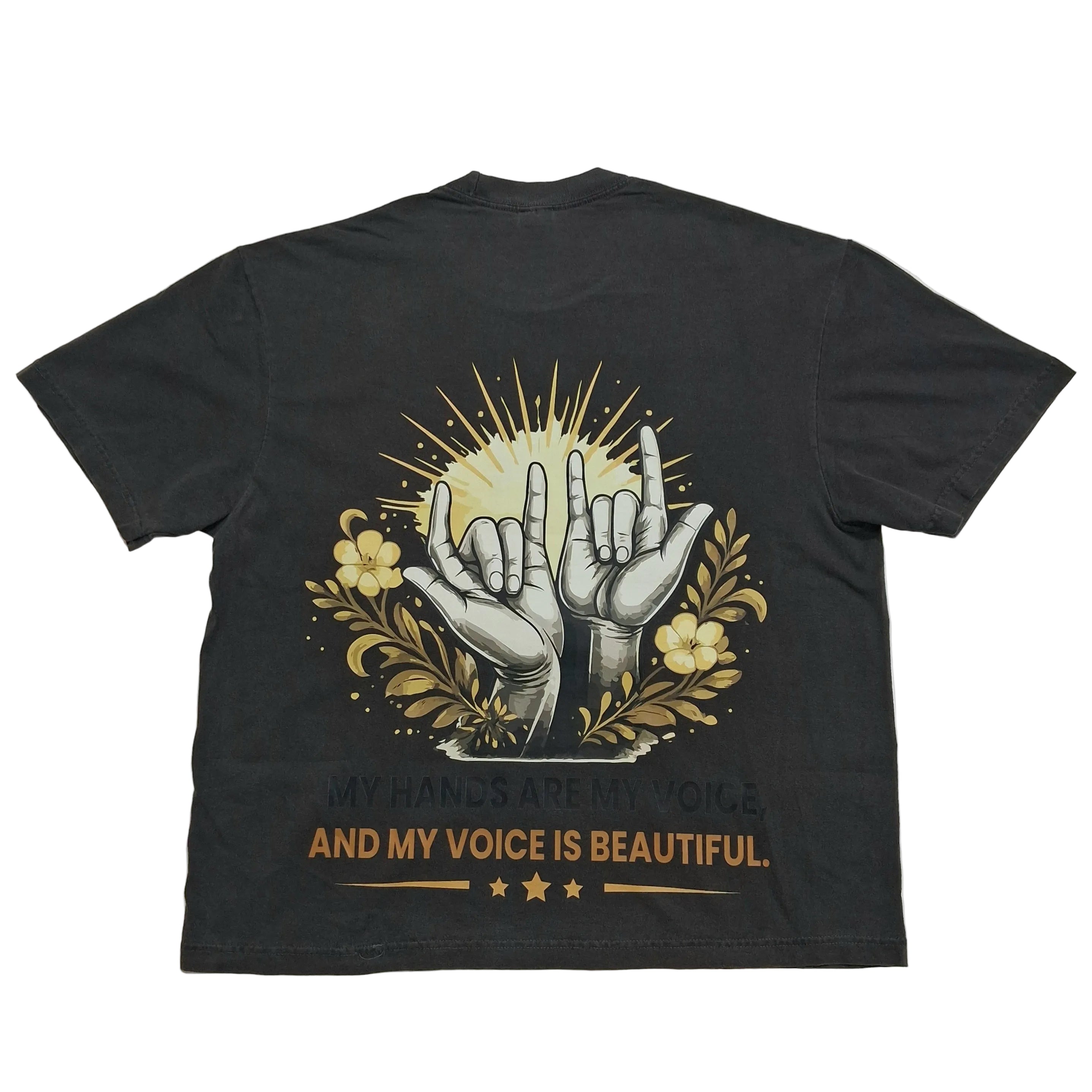 A black short-sleeve t-shirt laid flat against a light gray background, showing the back side with a graphic print of two hands making the shaka sign, surrounded by yellow flowers and rays, with the text 'MY HANDS ARE MY VOICE AND MY VOICE IS BEAUTIFUL.'.