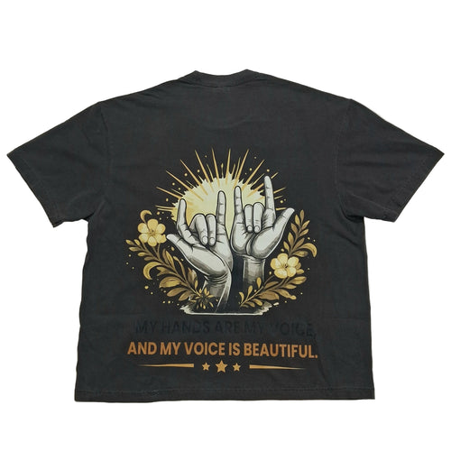 A black short-sleeve t-shirt laid flat against a light gray background, showing the back side with a graphic print of two hands making the shaka sign, surrounded by yellow flowers and rays, with the text 'MY HANDS ARE MY VOICE AND MY VOICE IS BEAUTIFUL.'.