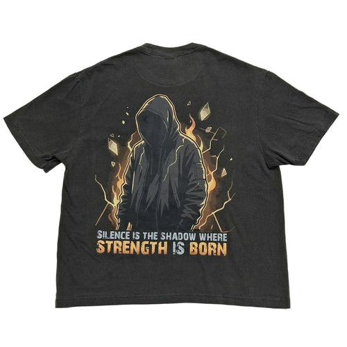 A dark charcoal gray graphic t-shirt laid flat on a light gray background. The Back of the shirt features a graphic of a hooded figure surrounded by orange and yellow flames, with the text 'SILENCE IS THE SHADOW WHERE STRENGTH IS BORN' printed below in white and orange.