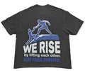 A dark charcoal gray t-shirt showing the back side, which features a graphic of two blue figures lifting each other up. Below the graphic, the text reads 'WE RISE by lifting each other.